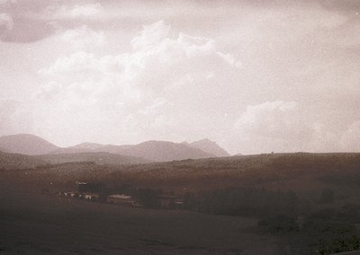 Vintage sepia-toned landscape featuring rolling hills under dramatic, textured clouds. Distant structures suggest rural settl...