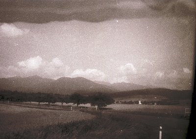 Vintage sepia-toned landscape featuring rolling hills, sparse trees, and a winding road under a dramatic sky. Low-contrast gr...