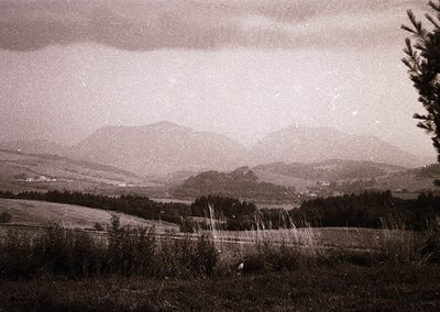 Vintage sepia-toned landscape featuring rolling hills, dense forests, and distant mountains under a moody sky. Foreground sho...
