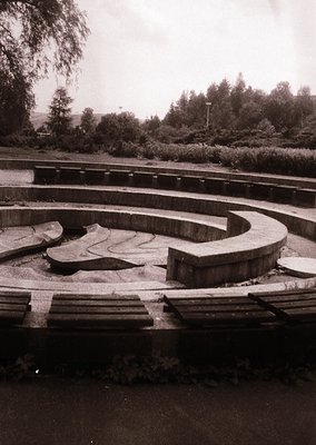 Concrete maze pathway in a landscaped park, featuring concentric loops with worn edges and overgrown vegetation. Mid-20th cen...