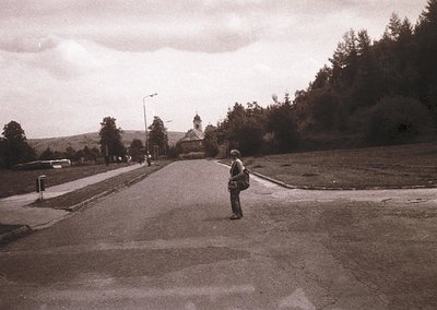 Black-and-white street scene featuring a lone pedestrian in a wide-brimmed hat and dark coat, walking toward a distant church...
