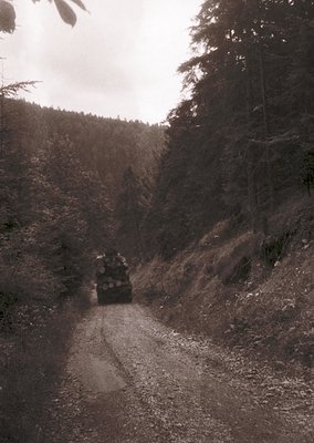 Black-and-white vintage photo of a rugged dirt road winding through a forested mountain pass. A military truck with camouflag...