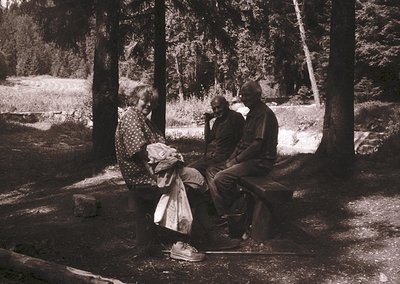 Three individuals seated on a stone bench in a forested area, mid-20th century. The woman wears a patterned blouse and light-...