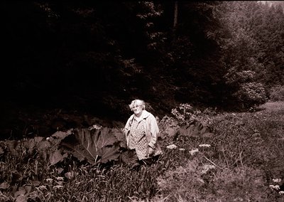 Black-and-white portrait of an elderly woman standing on a forested path, surrounded by dense foliage and large leaves. She w...