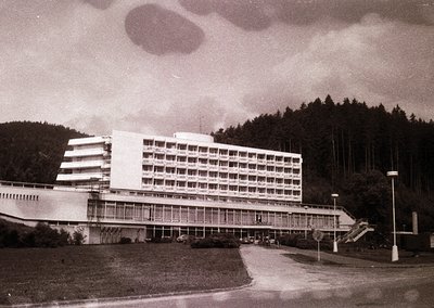 Mid-century Brutalist-style hotel with curved concrete balconies and extensive glass paneling, set against a forested hillsid...