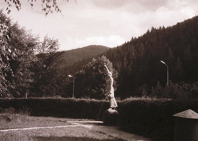 Black-and-white street scene featuring a lone figure casting a fishing line into a shallow riverbed, framed by dense foliage ...