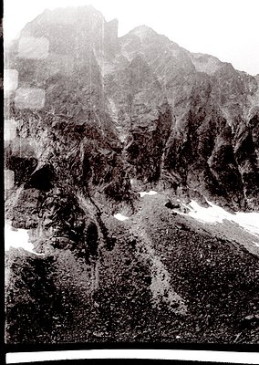 Black-and-white aerial view of rugged alpine terrain with jagged peaks, steep slopes, and scattered snow patches. Likely capt...