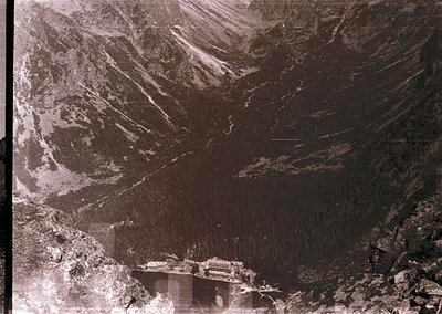 Aerial view of a mountainous region with a prominent dam and reservoir. The dam appears concrete, spanning a valley with fore...