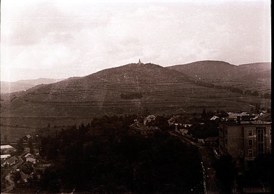 Vintage sepia-toned aerial view of a small European town nestled between terraced hills and dense forest. Prominent church sp...
