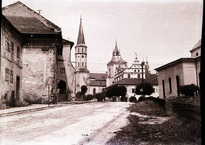 Historic European village street with a mix of Gothic and Baroque architecture. Prominent church tower with a spire and adjac...