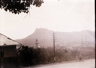 Black-and-white street scene featuring a hilltop fortress and Ottoman-era minarets in the background. Two pedestrians walk al...