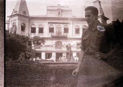 Vintage sepia-toned photo of a grand colonial-era building with arched balconies, central tower, and lush greenery. Uniformed...