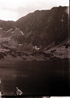 Mid-20th century black-and-white alpine lake scene with rugged peaks. A lone figure in outdoor gear sits by the water’s edge,...