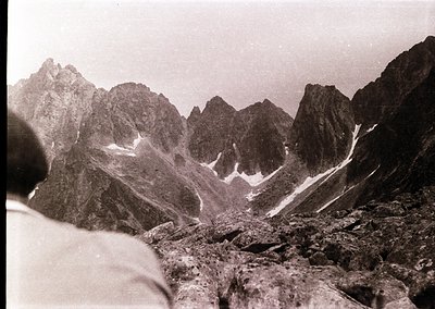 Early 20th-century black-and-white alpine landscape showing jagged peaks and rocky terrain. Partial figure in foreground sugg...