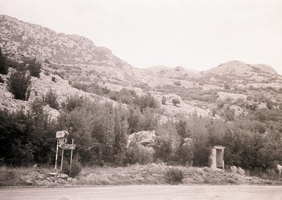 Vintage black-and-white roadside scene with a directional signpost reading "Peshtera" and a public toilet structure. Rugged m...