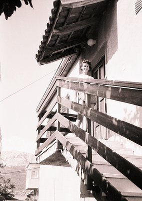 A woman leans from a wooden balcony railing, mid-20th century. Wearing a sleeveless dress, she gazes upward toward a sloped r...