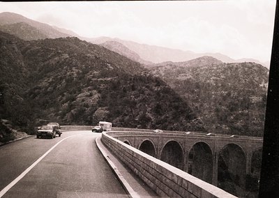 Mid-20th century highway bridge spanning lush, forested mountains. Concrete viaduct supports multiple lanes with vintage cars...