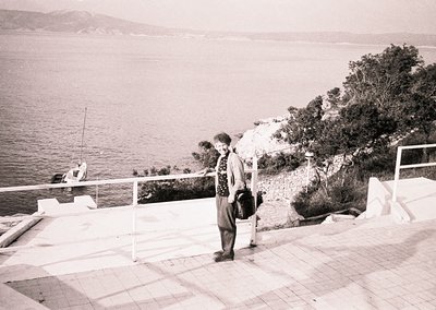 Mid-20th century seaside promenade with a lone woman in patterned dress and headscarf, holding a bag. White balustrade overlo...