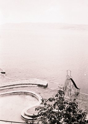 Vintage black-and-white seaside pool with curved concrete edges and a diving tower in foreground. Overcast sky and calm water...