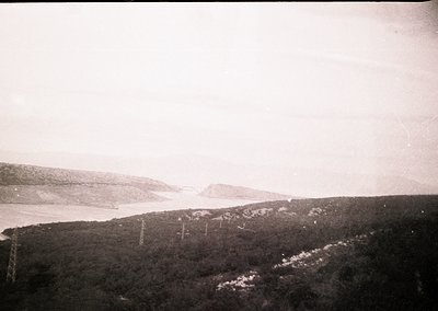 Vintage aerial view of coastal terrain with dense forest and rocky coastline. Power lines and transmission towers stretch acr...