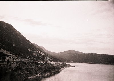 Vintage black-and-white lakeside scene with rugged mountain range framing a serene body of water. Dense forest clings to hill...
