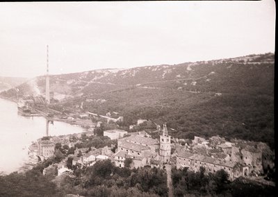 Black-and-white aerial view of a coastal settlement with a prominent church steeple and industrial tower. Dense foliage frame...