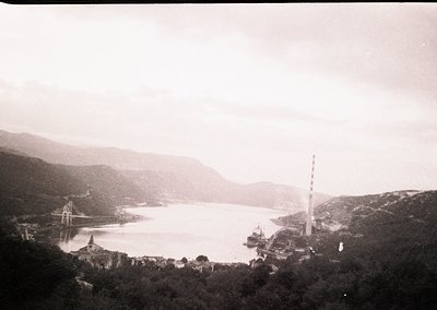 Vintage black-and-white coastal scene featuring a prominent radio tower on a hillside, a suspension bridge spanning a calm ba...