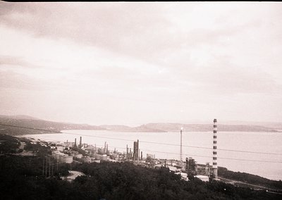 Black-and-white aerial view of an industrial coastal facility, likely a refinery or petrochemical plant, with multiple tall c...