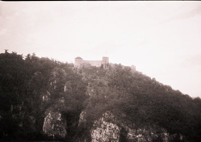 Medieval fortress perched atop rugged cliffs, surrounded by dense forest. Stone walls and towers suggest defensive architectu...
