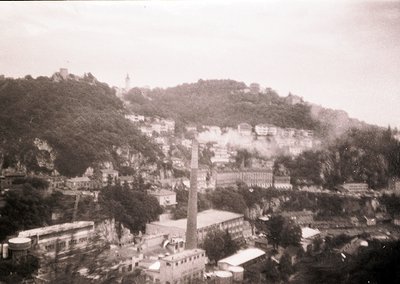 Vintage black-and-white aerial view of a hilly coastal town with dense, irregularly built structures. Prominent industrial ch...