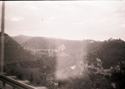 Vintage black-and-white view of a suspension bridge spanning a forested valley, framed by a window. Mid-20th century industri...