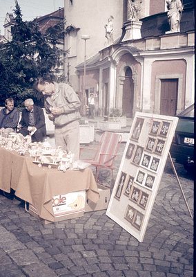 Street-side vintage market in a European town square, likely 1960s–70s. Three men examine ceramic figurines and framed religi...