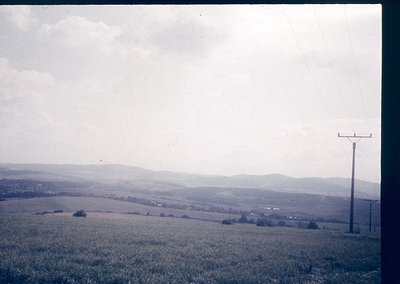 Vintage landscape shot of rolling hills and open fields under overcast skies. A lone utility pole with crossbar stands in the...