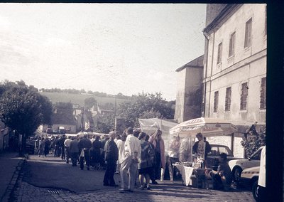 Vintage street market scene with vintage cars, likely 1960s–1970s. Crowd browsing stalls under makeshift canopies; rural Euro...