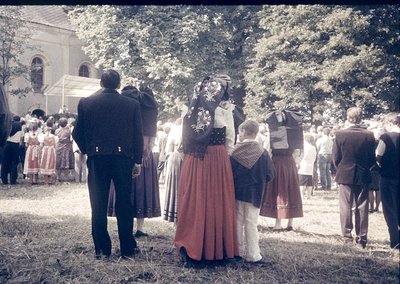 Vintage outdoor gathering featuring traditional folk attire—men in dark suits, women in embroidered skirts and blouses with f...