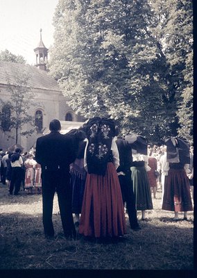 Vintage outdoor gathering featuring traditional folk attire—men in dark suits with white shirts, women in embroidered dresses...