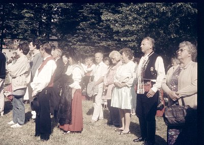 Group of adults in traditional folkwear, likely Eastern European, gathered outdoors in a wooded area. Men wear embroidered ve...
