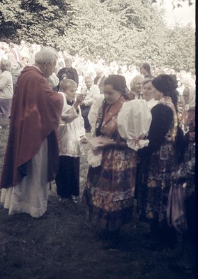Vintage outdoor gathering featuring a priest in liturgical vestments blessing a woman in traditional folk attire, surrounded ...