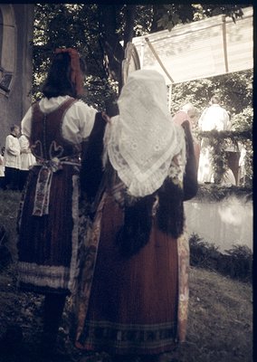 Traditional folk costumes featuring embroidered aprons and lace headscarves, likely Eastern European. Wooden structure and gr...