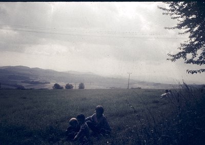 Vintage black-and-white rural landscape featuring three seated figures in grassy field. Rolling hills and power lines stretch...