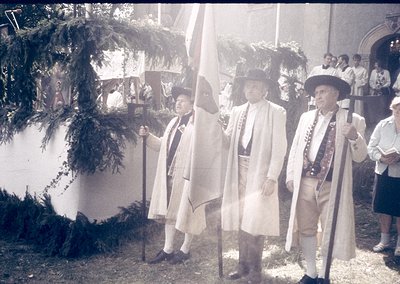 Four men in 18th-century military uniforms (tricorne hats, white coats, sashes) pose outdoors near a building with arched ent...