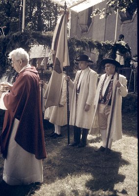 Traditional folk costumes and ceremonial flag procession in rural Europe, likely 1960s–1980s. Three men in white linen robes ...
