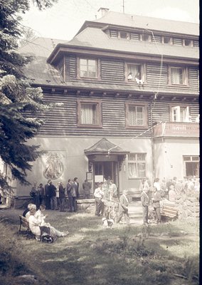 Vintage alpine lodge with log-clad facade, likely 1920s-1940s. Crowd gathers on steps and lawn, suggesting social or communal...