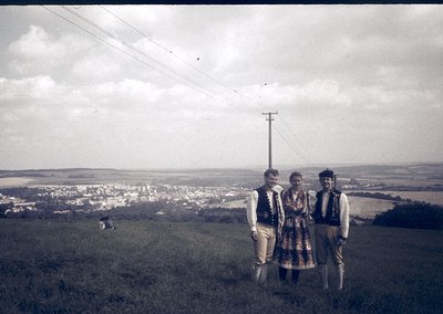 Three individuals in traditional folkwear pose on a grassy hilltop, overlooking a rural settlement. The man on the left wears...