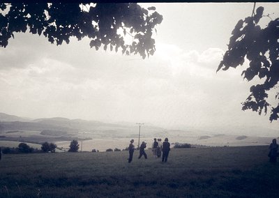 Vintage black-and-white photo of a group of hikers ascending a grassy hillside, framed by tree branches. Rolling countryside ...