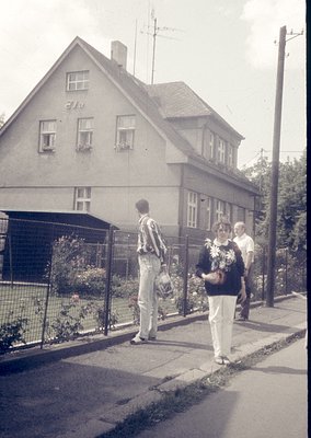Two men and a woman in 1970s streetwear stand outside a two-story, gabled house with whitewashed walls and wooden-framed wind...