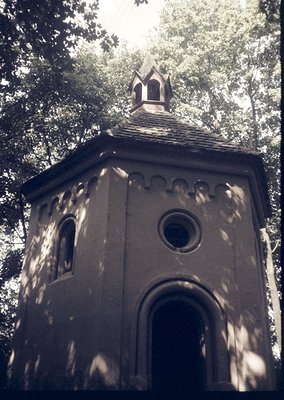 Historic stone chapel with pointed bell tower and arched windows, set in a wooded area. Architectural details include a small...