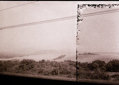 Vintage sepia-toned coastal landscape with dense greenery framing a distant shoreline and water body. Damaged corner reveals ...