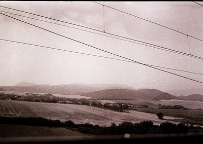 Mid-20th century rural landscape with rolling hills and open fields, framed by power lines. Vintage black-and-white grain add...