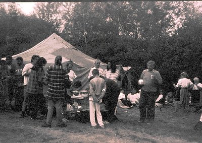 Vintage outdoor gathering with a group of people around a makeshift grill under a large canvas tent. Mid-20th century clothin...